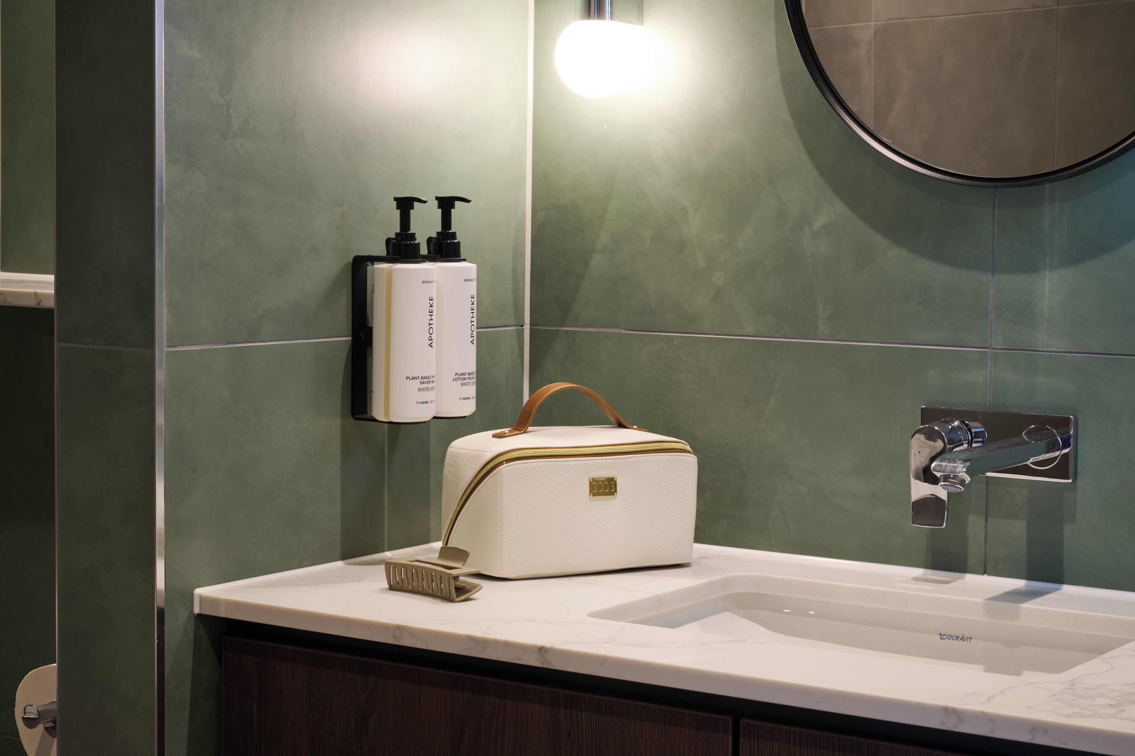 A bathroom with a white sink, a handheld brush, a white toiletry bag, and two bottles of lotion on the wall.