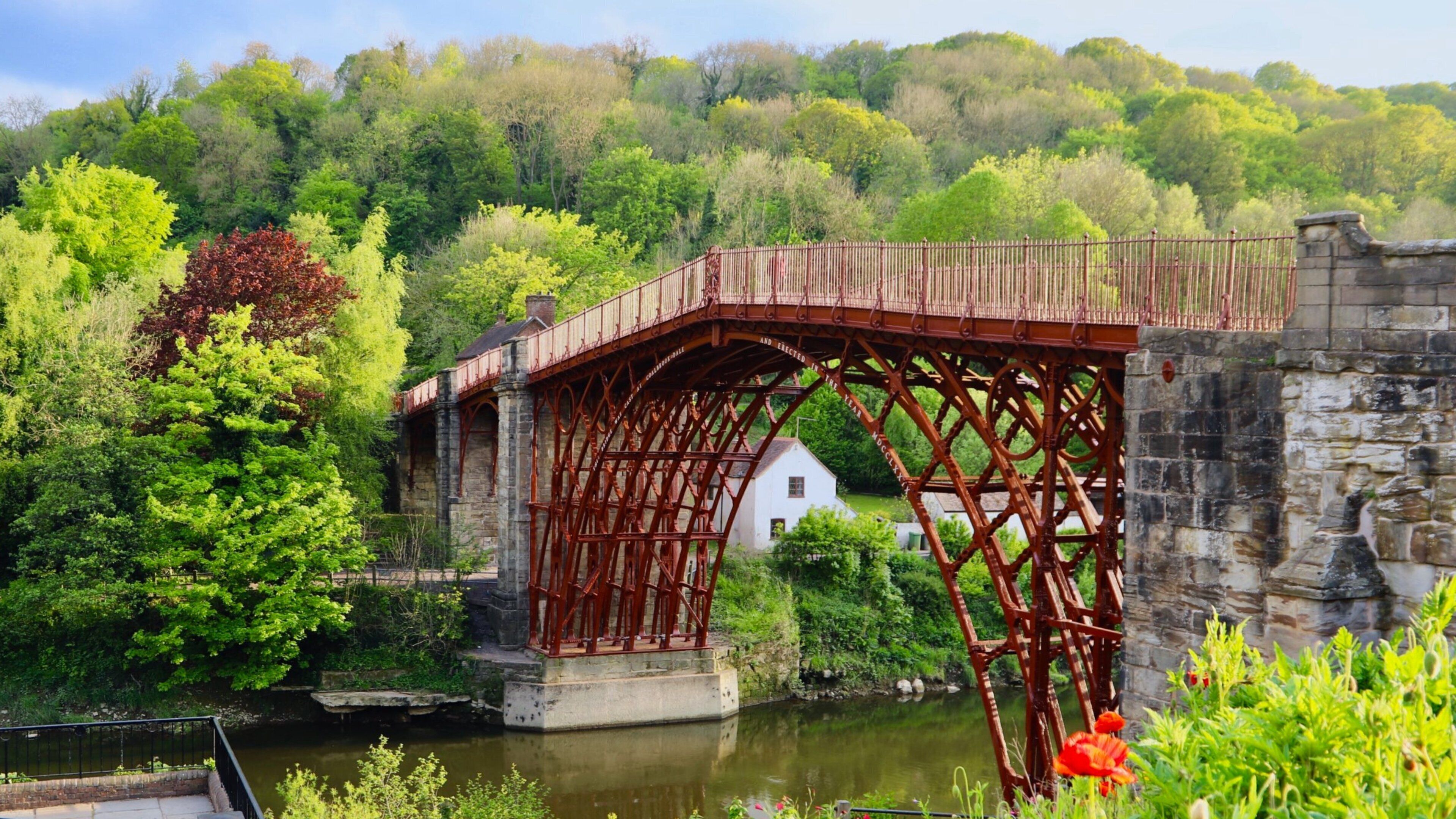 Un pont de pierre historique doté d’une structure métallique, un bâtiment blanc en arrière-plan et une végétation luxuriante bordant une étendue d’eau.