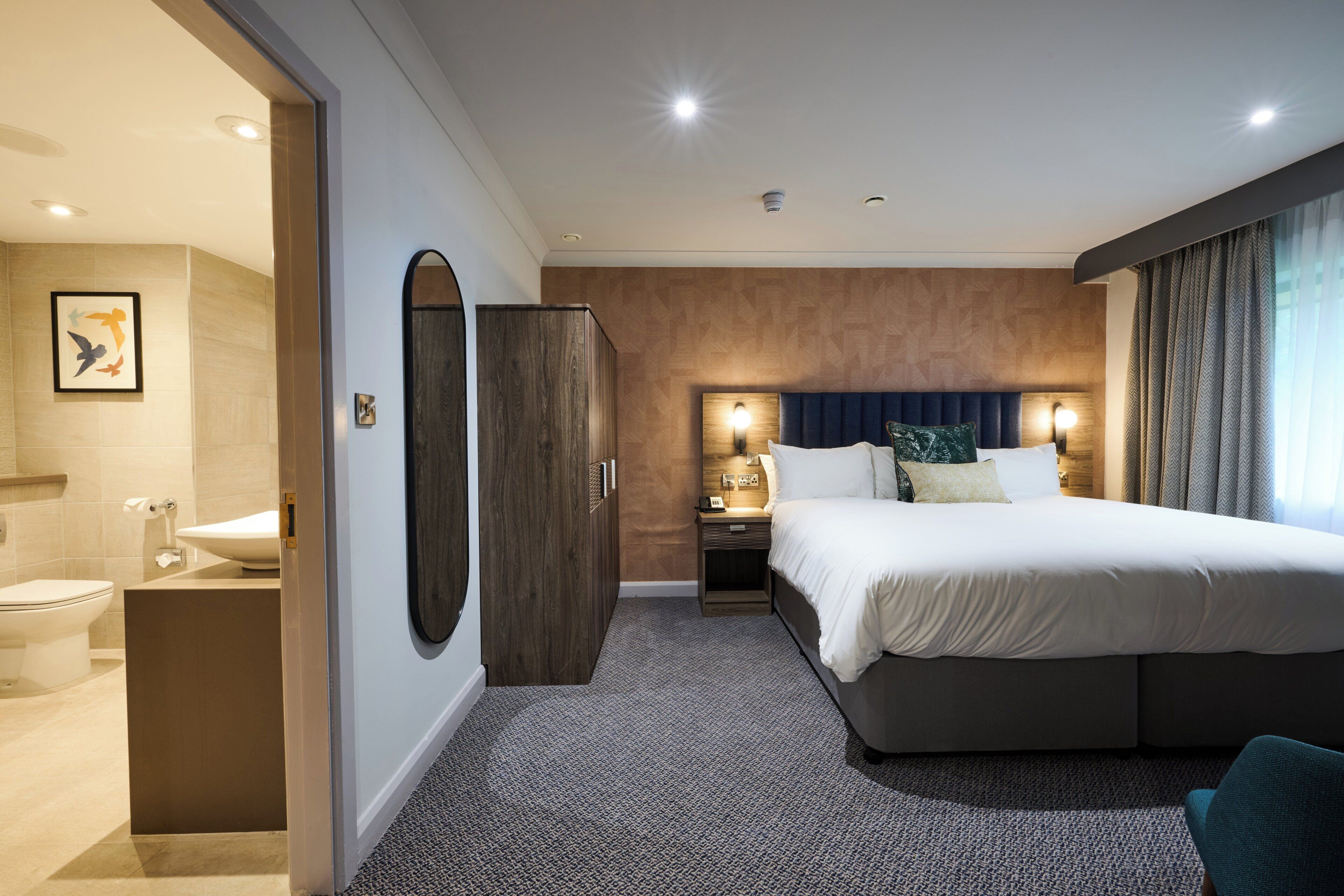 A hotel room with a large bed, a bedside table, a nightstand, a mirror, and a view of the bathroom.
