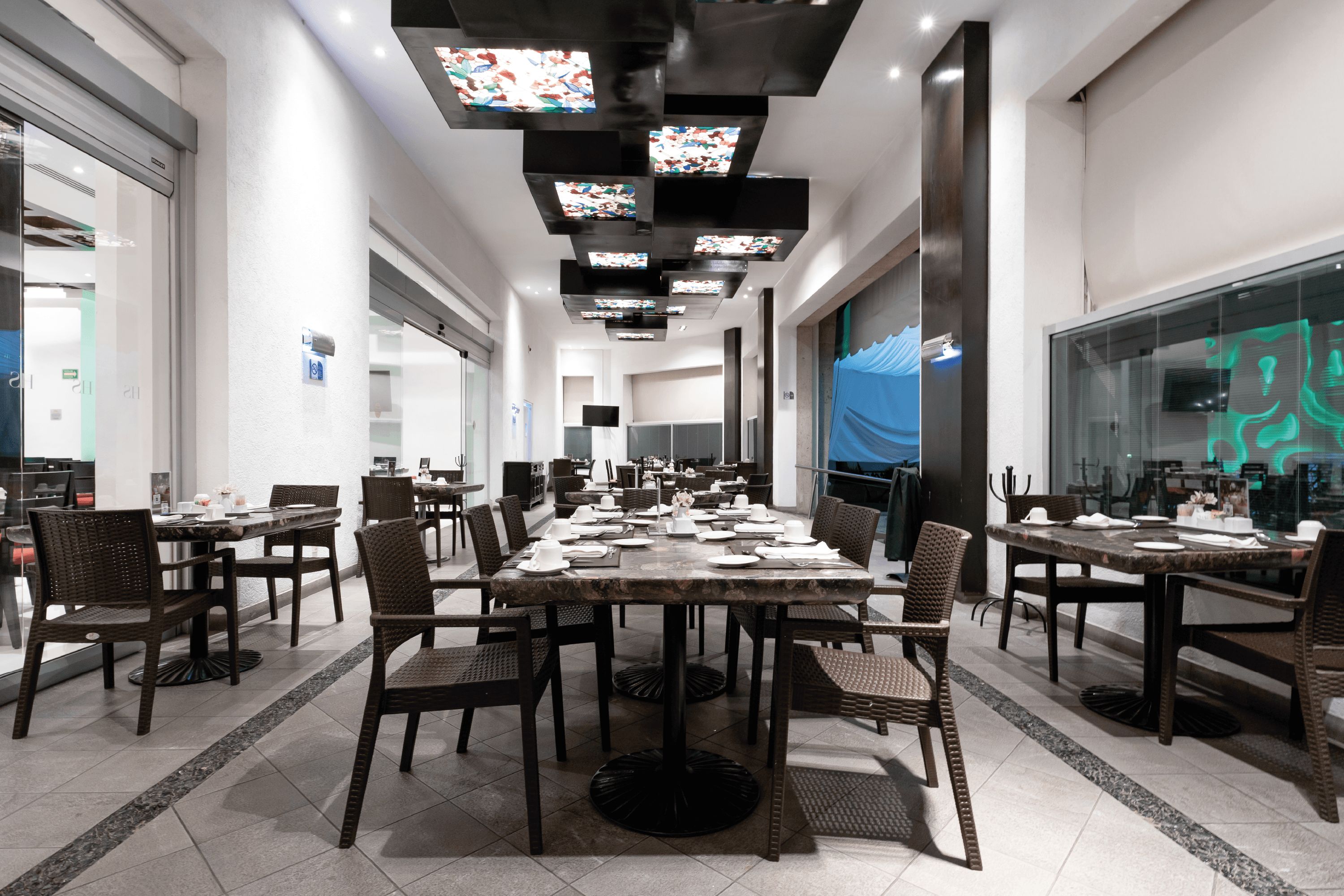 A spacious dining area with multiple tables and chairs, a modern ceiling design with recessed lighting, and a large window on the right side.