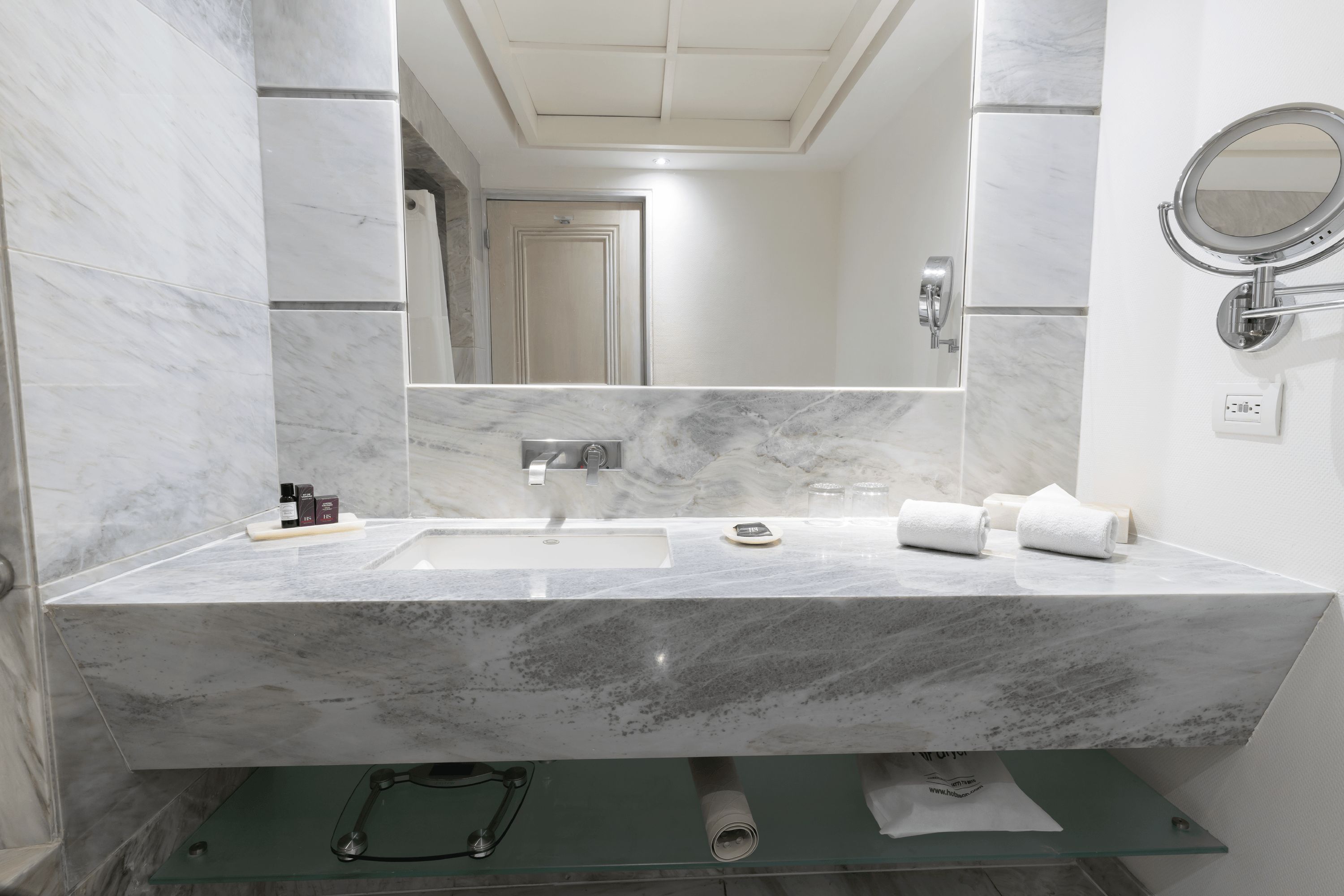 A bathroom with a marble sink, a mirror, a towel rack, and a glass shelf.