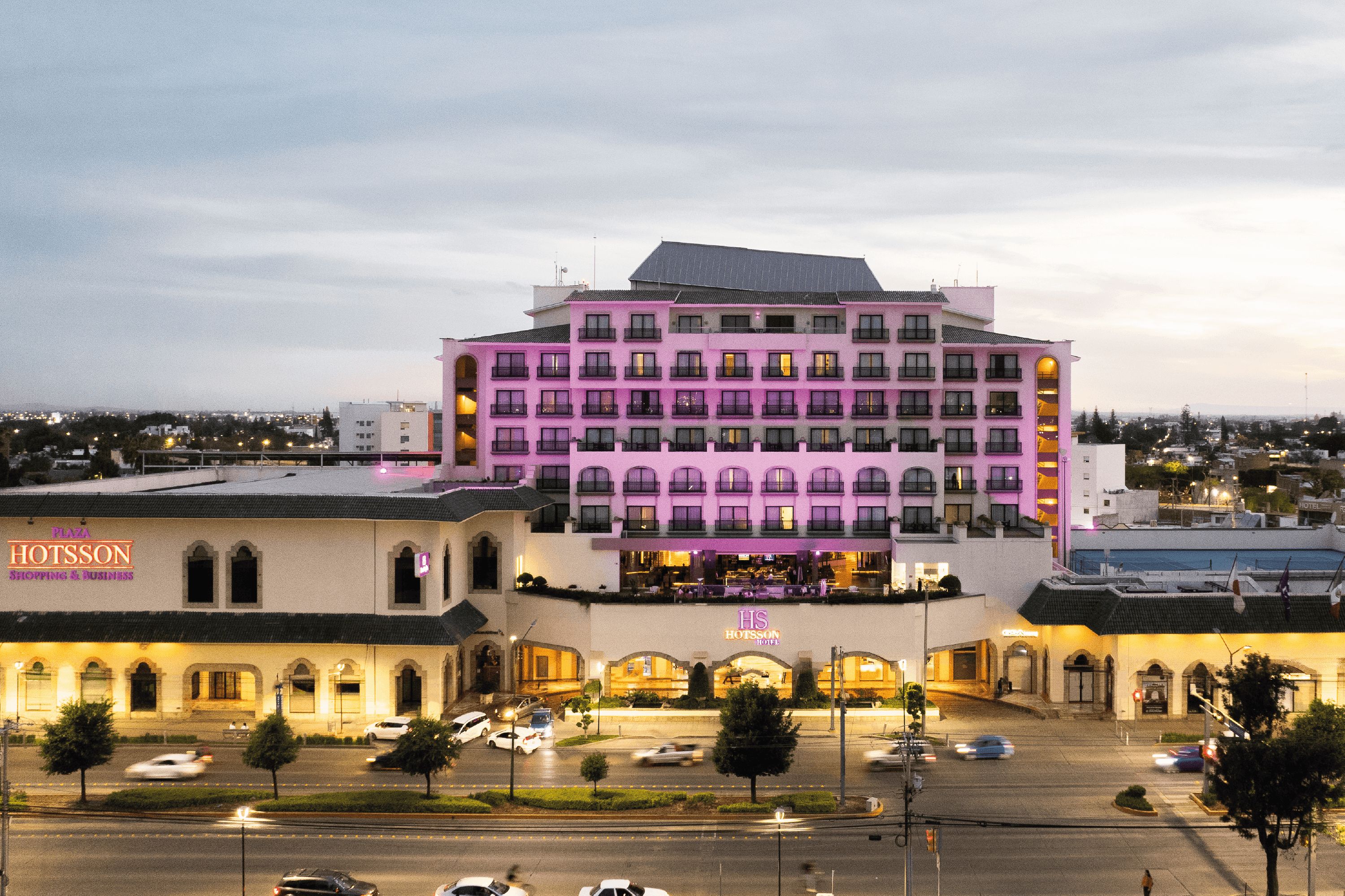 A multi-story hotel with a prominent pink facade, a ground-level shopping area, and a parking lot with cars.