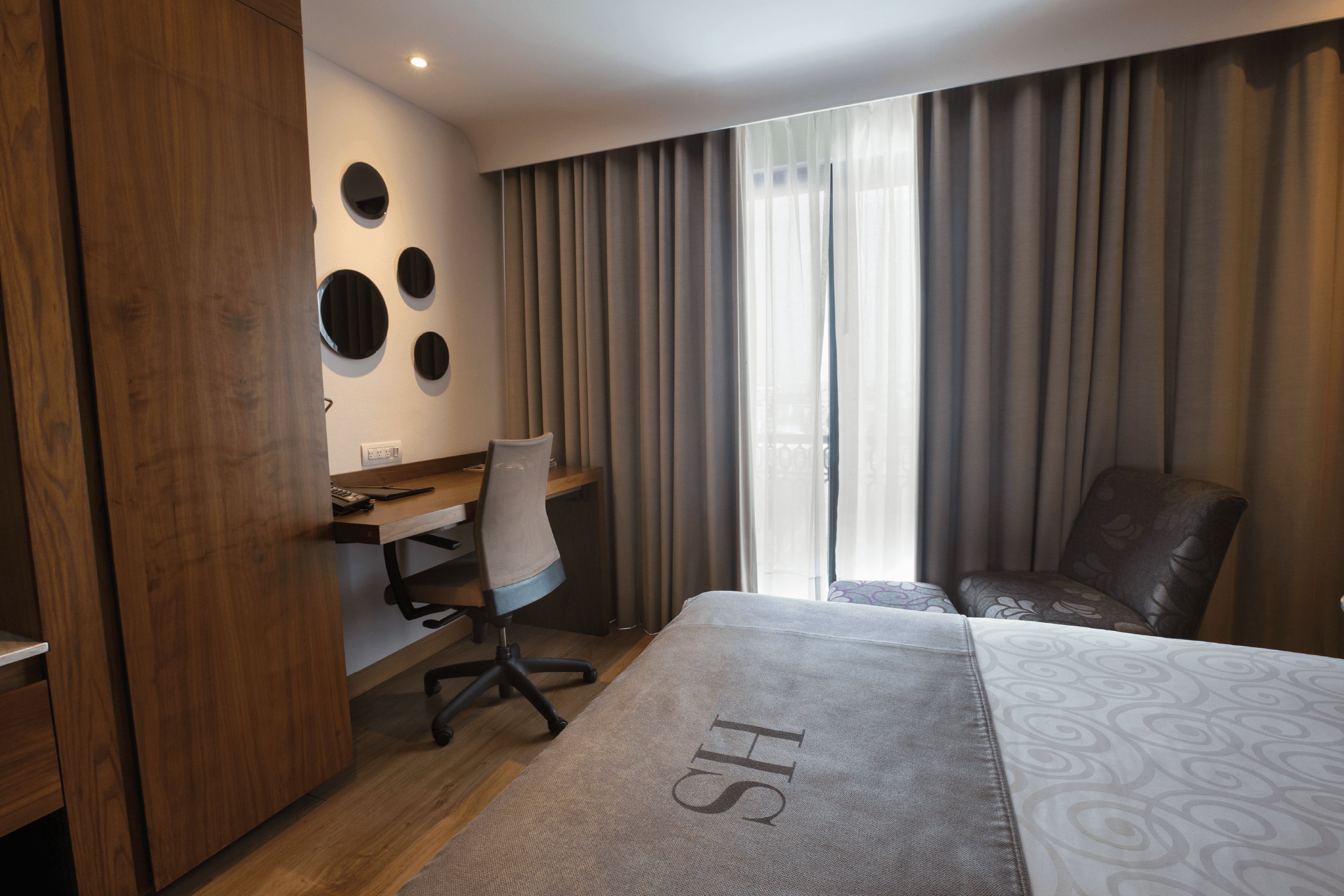 Habitación de hotel con cama, escritorio, silla, armario y ventana con cortinas.