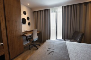 Habitación de hotel con cama, escritorio, silla, armario y ventana con cortinas.