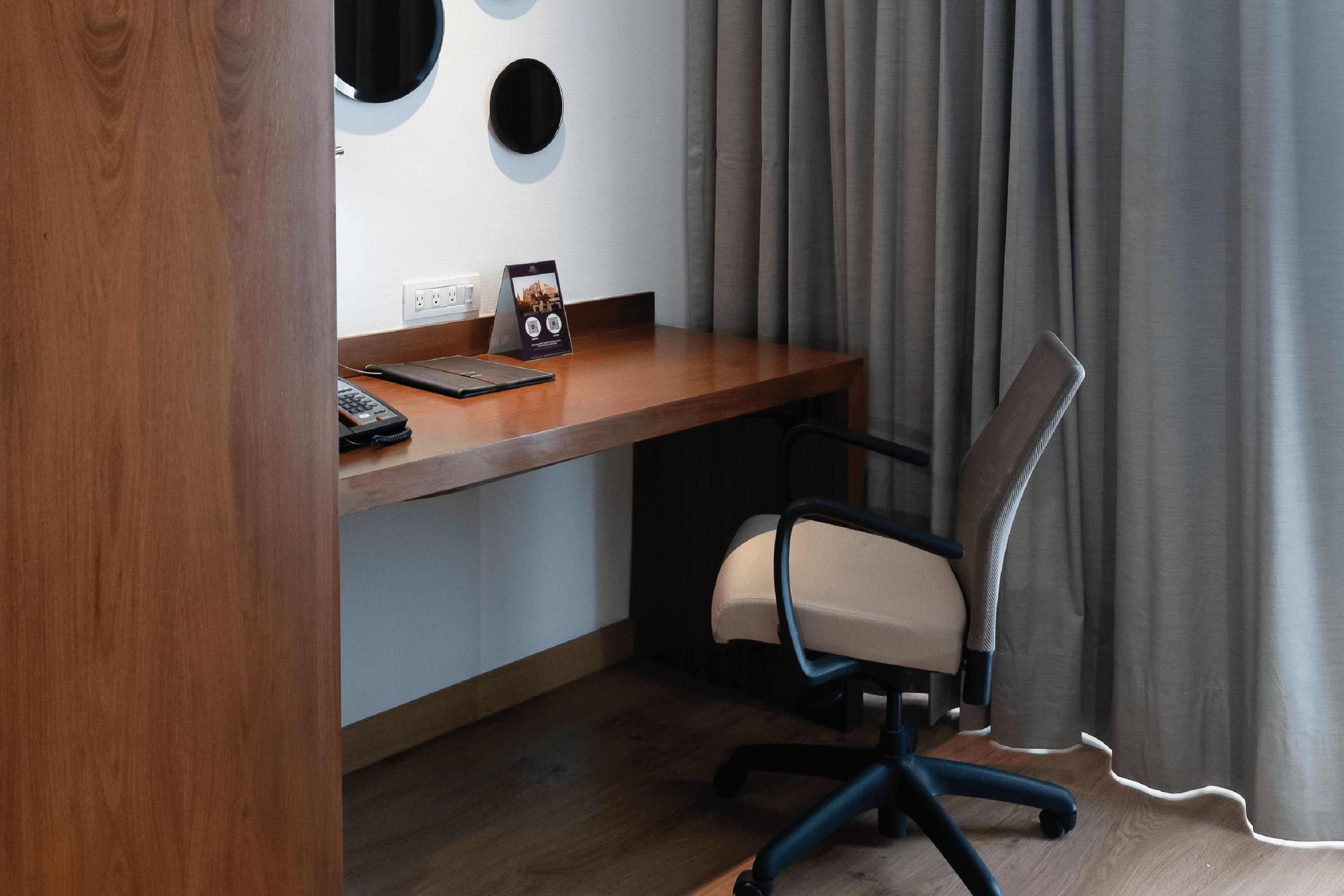 Una habitación de hotel con un escritorio de madera, una silla, un teléfono y un enchufe en la pared.
