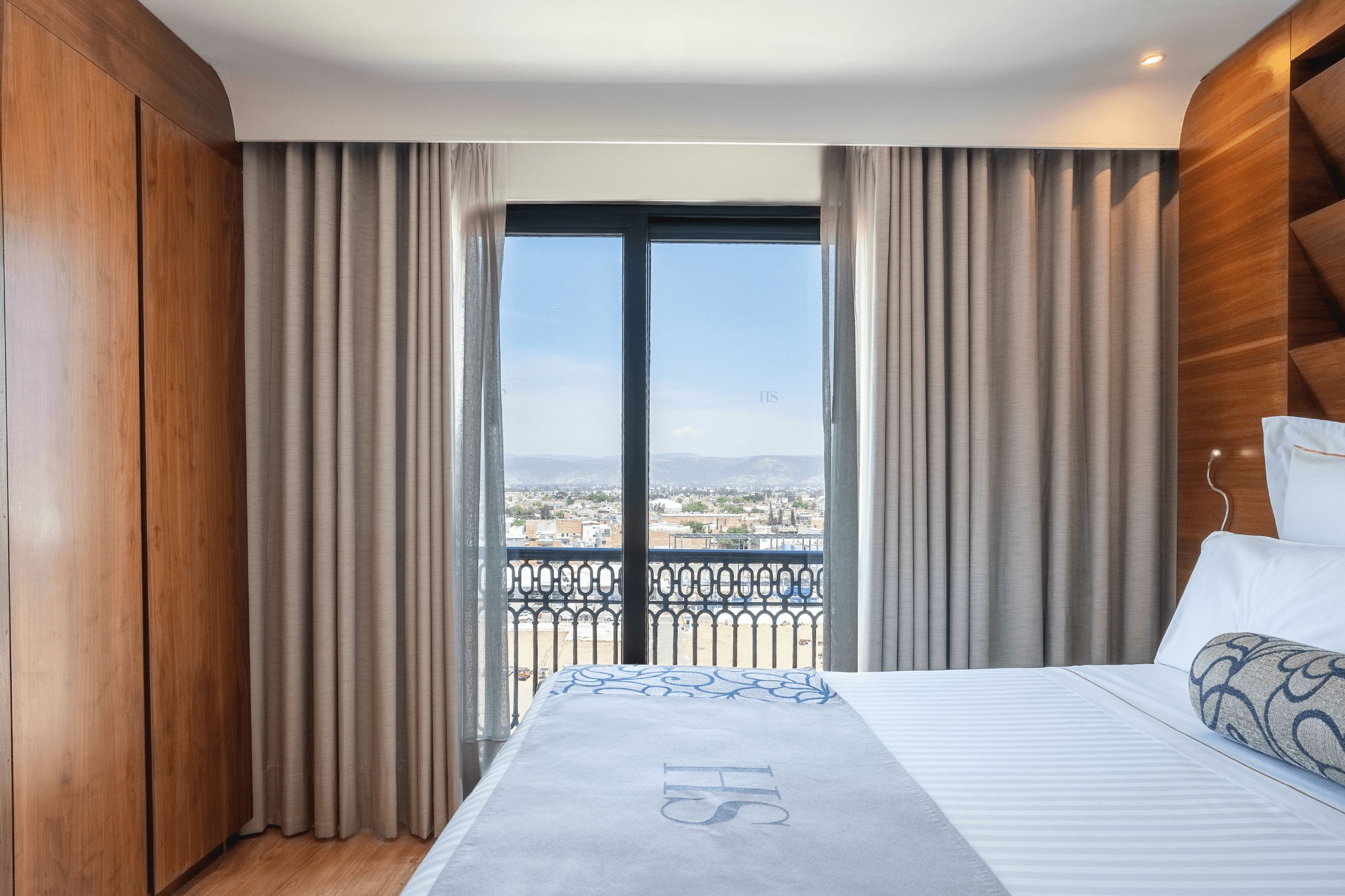 A hotel room with a large bed, a balcony with a view of the city, and wooden paneling on the walls.
