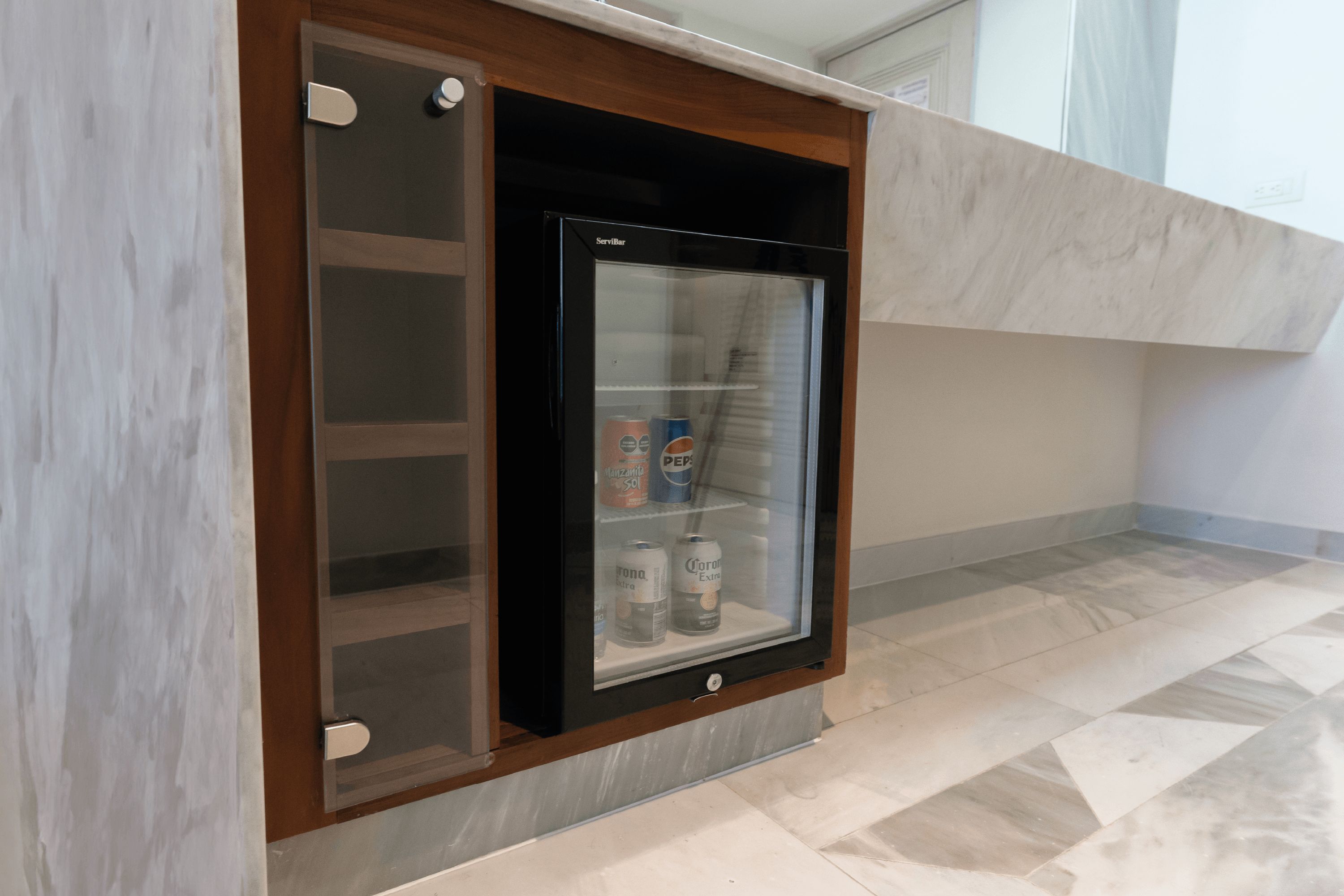Un mini-bar doté d’une porte en verre, contenant un réfrigérateur à boissons, une bouteille de Corona Extra et une canette de Pepsi.
