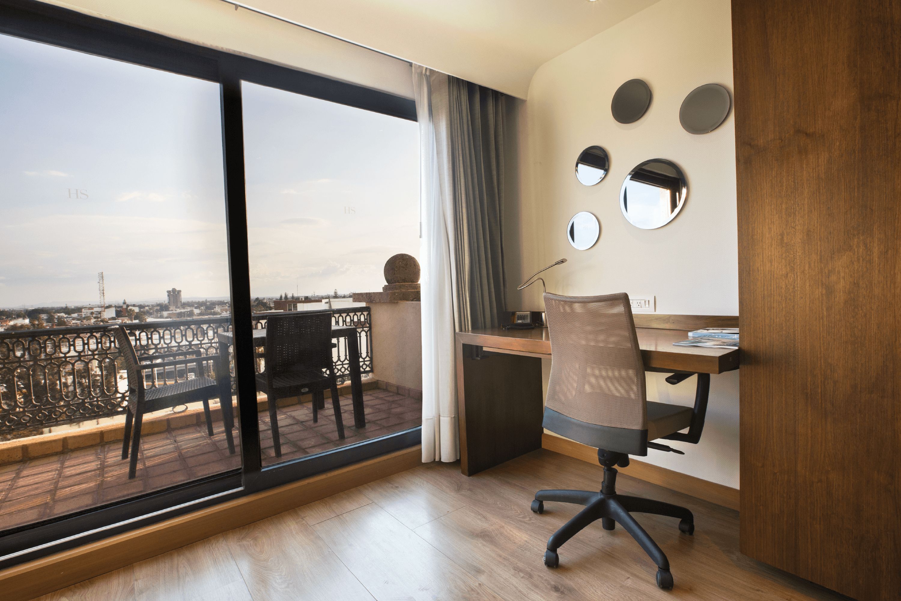 Un espace de bureau moderne doté d’une grande fenêtre, d’un bureau, d’une chaise et de miroirs décoratifs fixés au mur.