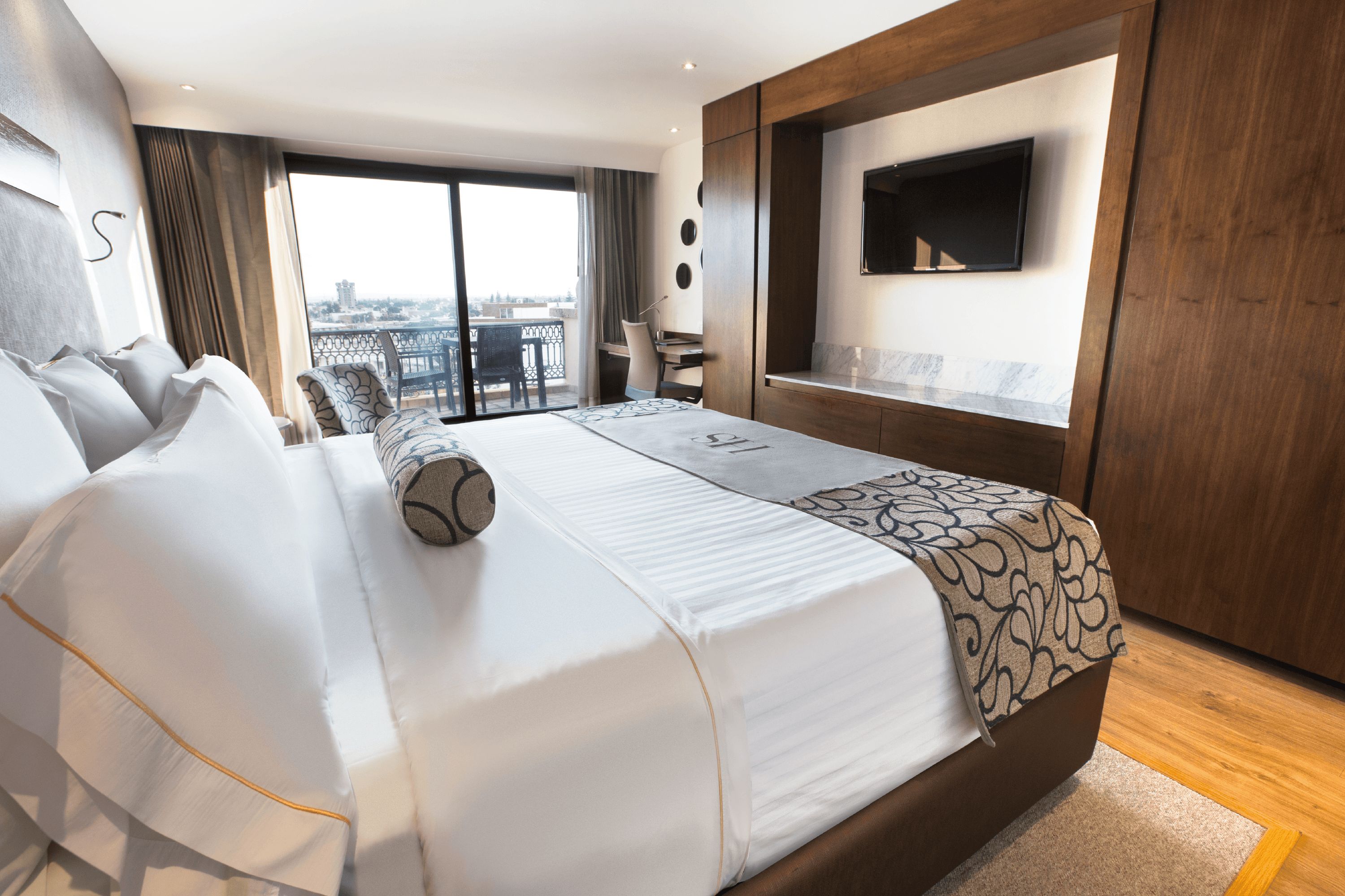 A hotel room with a large bed, a desk, a chair, a TV, and a view of the city through the window.