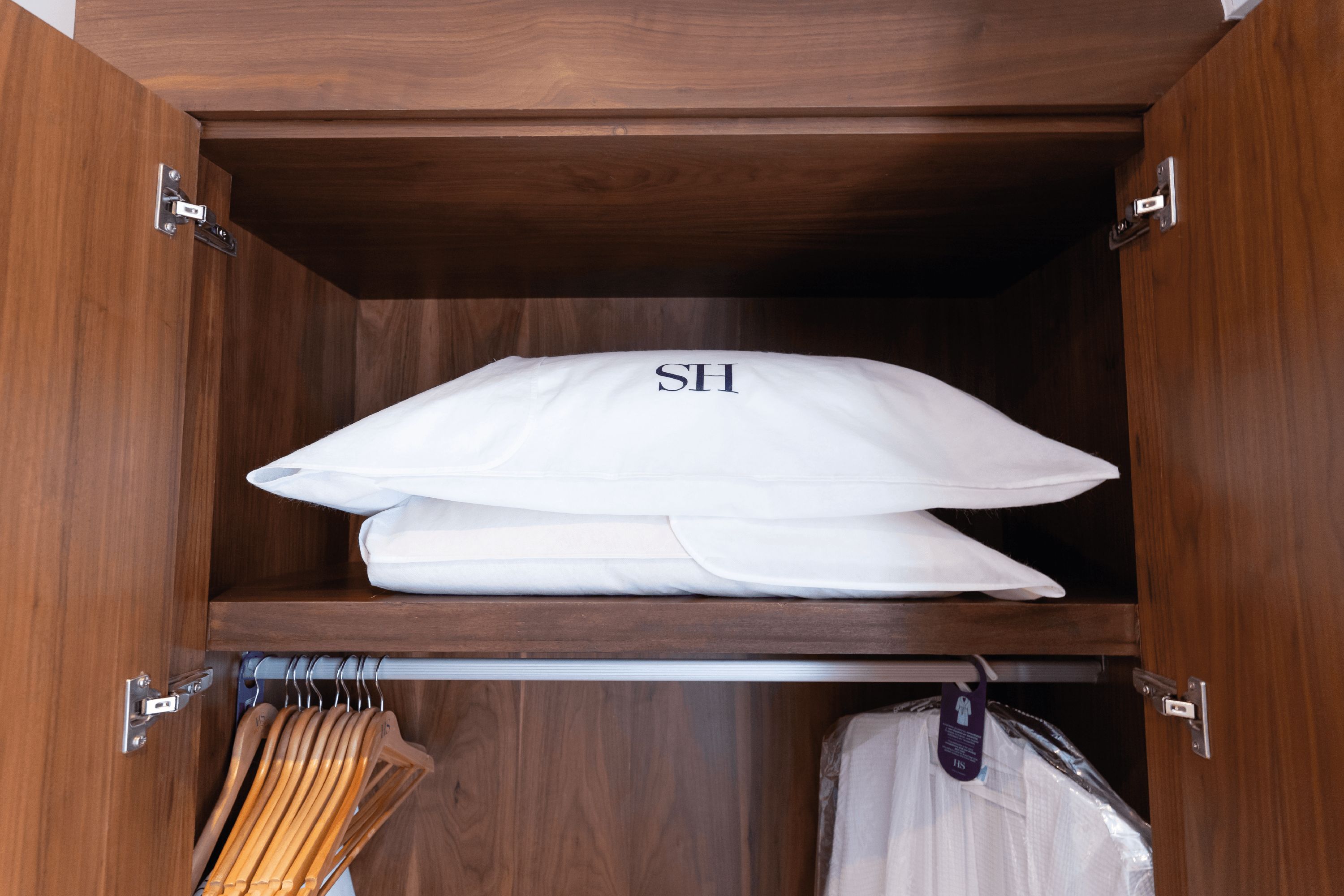 Ein Holzkleiderschrank mit Kleiderstange und einem gefalteten weißen Kopfkissen mit dem Aufdruck 'SH' darauf.
