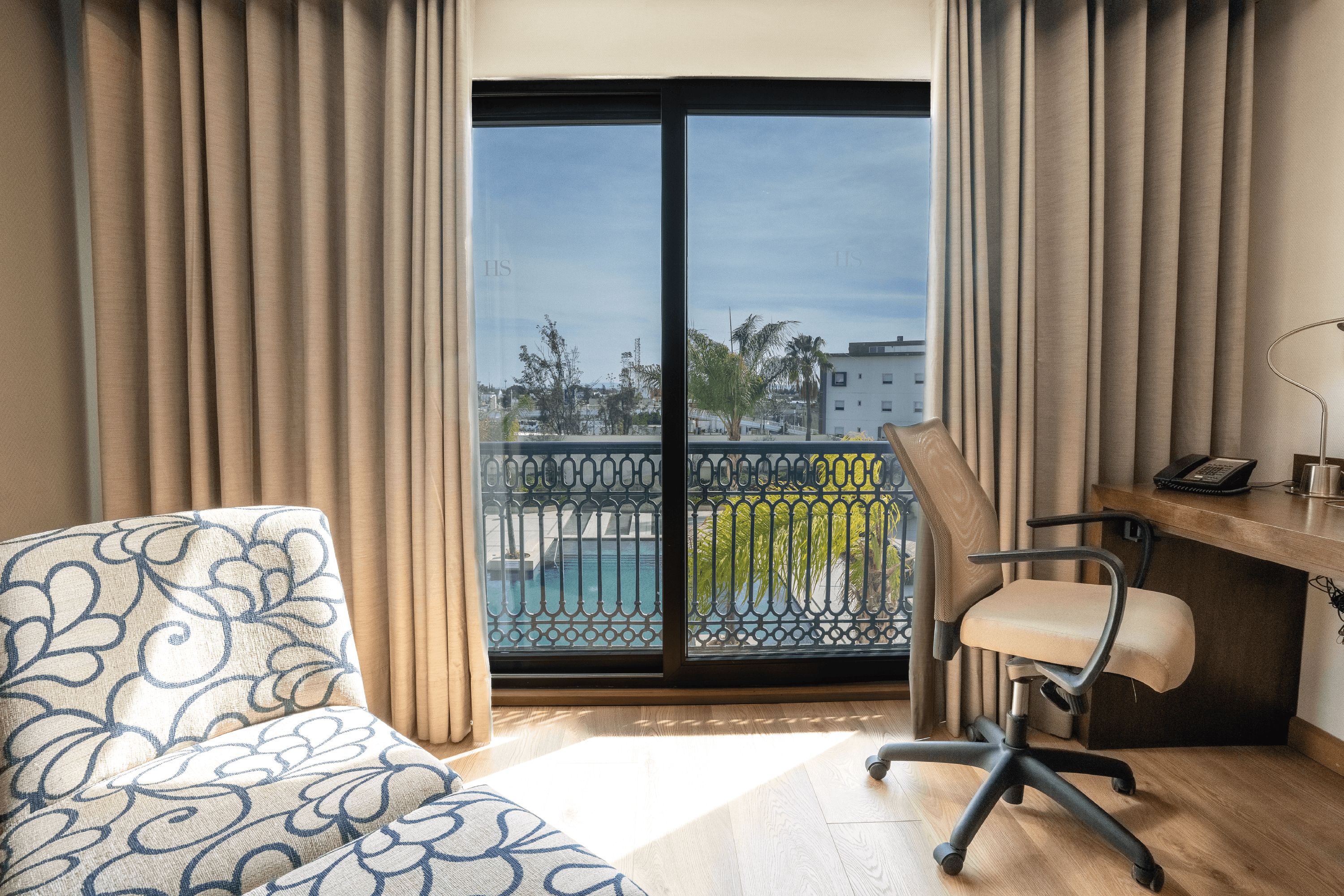 A hotel room with a balcony, a desk, a chair, and a patterned sofa.