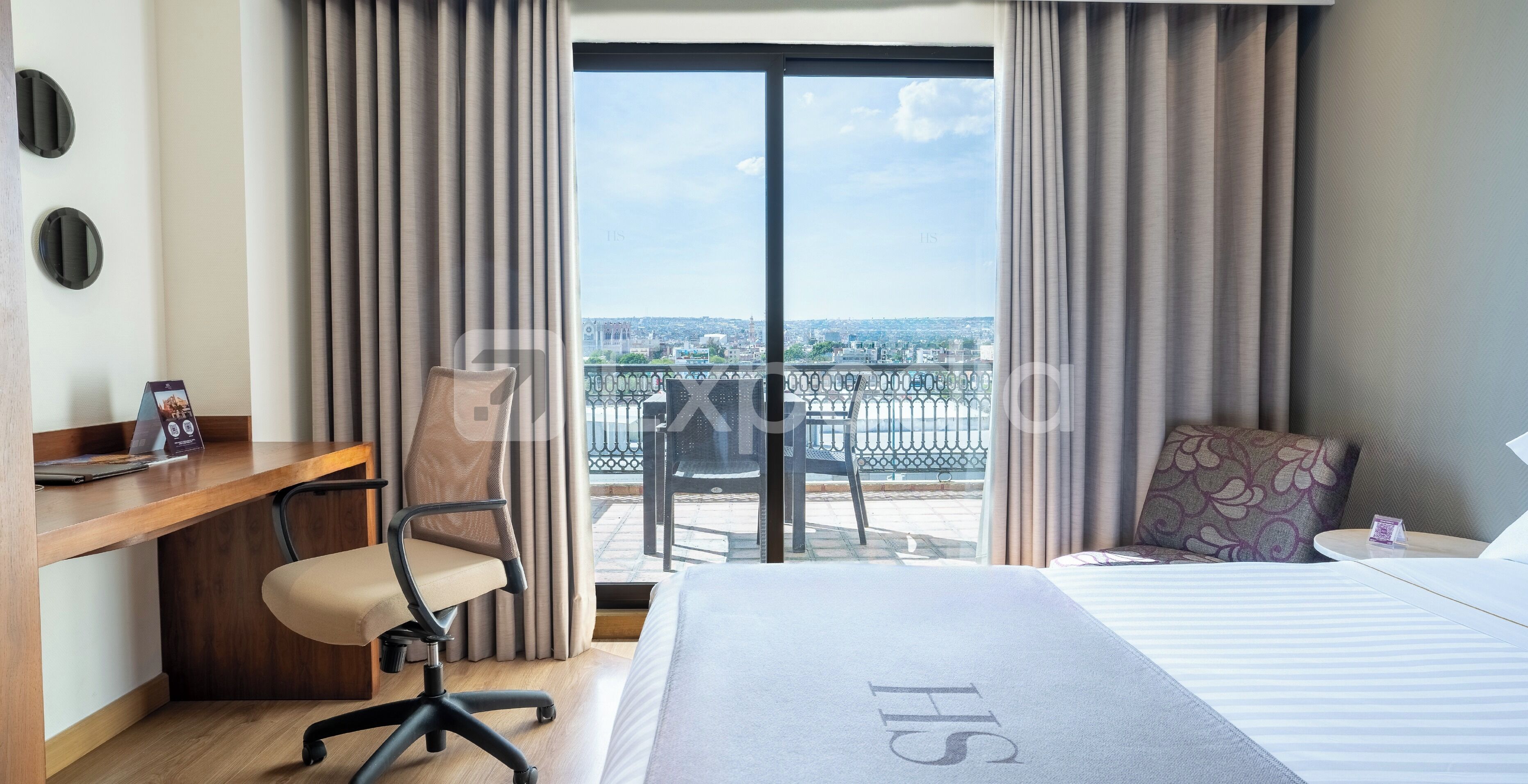 A hotel room with a bed, a desk, a chair, and a balcony with a city view.