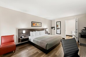 A modern hotel room with a large bed, a red armchair, a black office chair, a desk, and a bathroom visible through an open door.