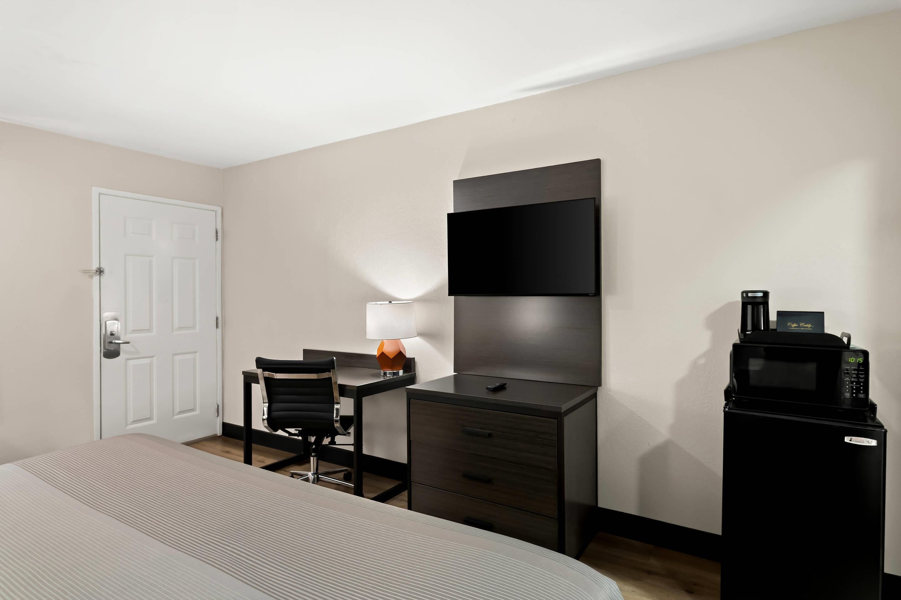 A hotel room with a bed, a desk with a chair, a small table with a lamp, a wardrobe, and a minibar.