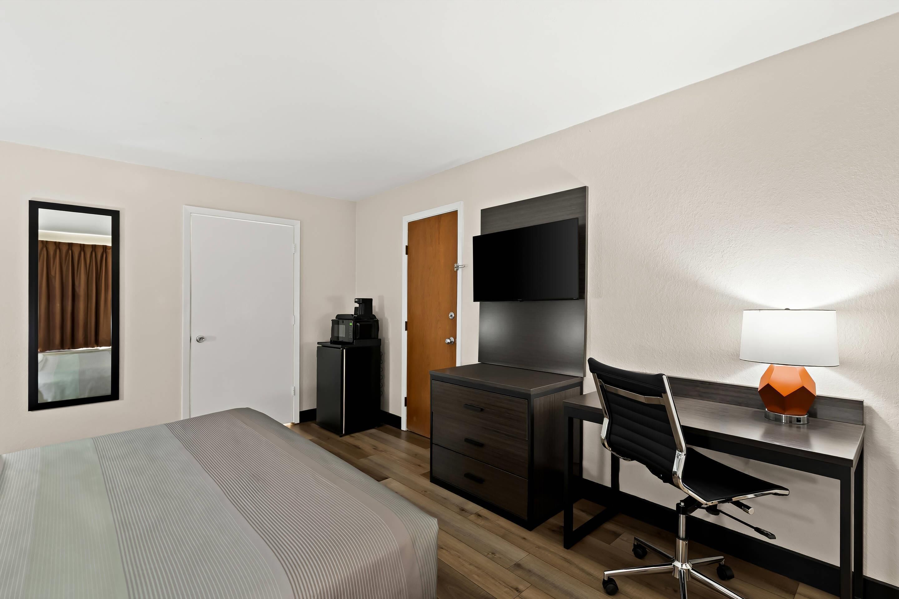 A hotel room with a bed, a desk with a chair, a TV, a lamp, and a wardrobe.