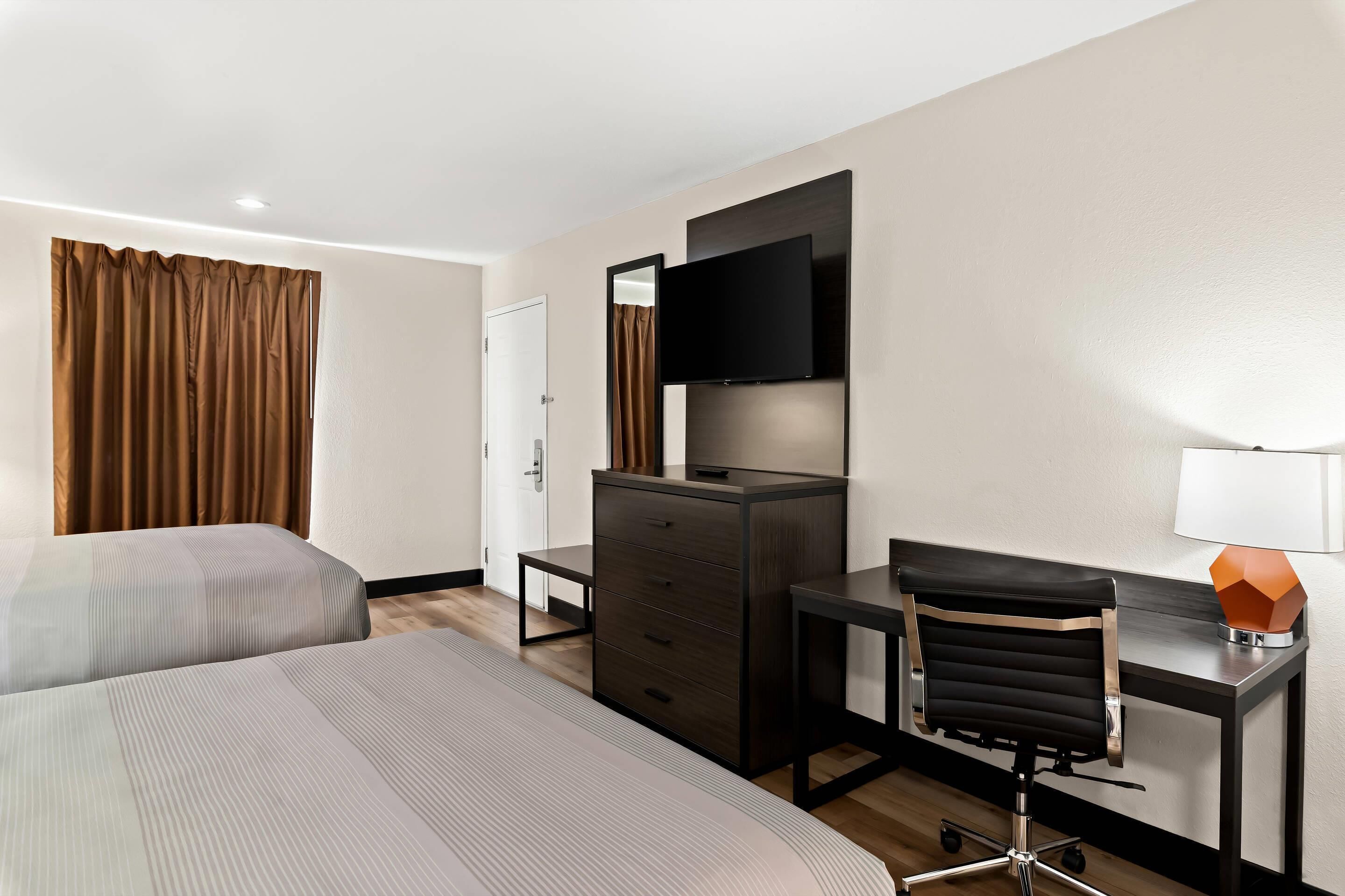 A hotel room with two beds, a desk with a chair, a dresser, a TV mounted on the wall, and a lamp on the desk.