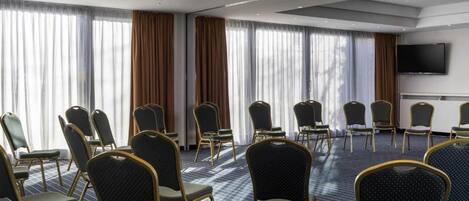 Une salle de conférence avec des rangées de chaises, de grandes fenêtres avec des rideaux et un téléviseur à écran plat fixé au mur.