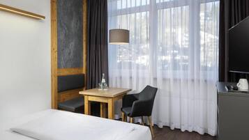 Habitación de hotel con cama, una mesa, dos sillas, televisión y un ventanal grande con cortinas.