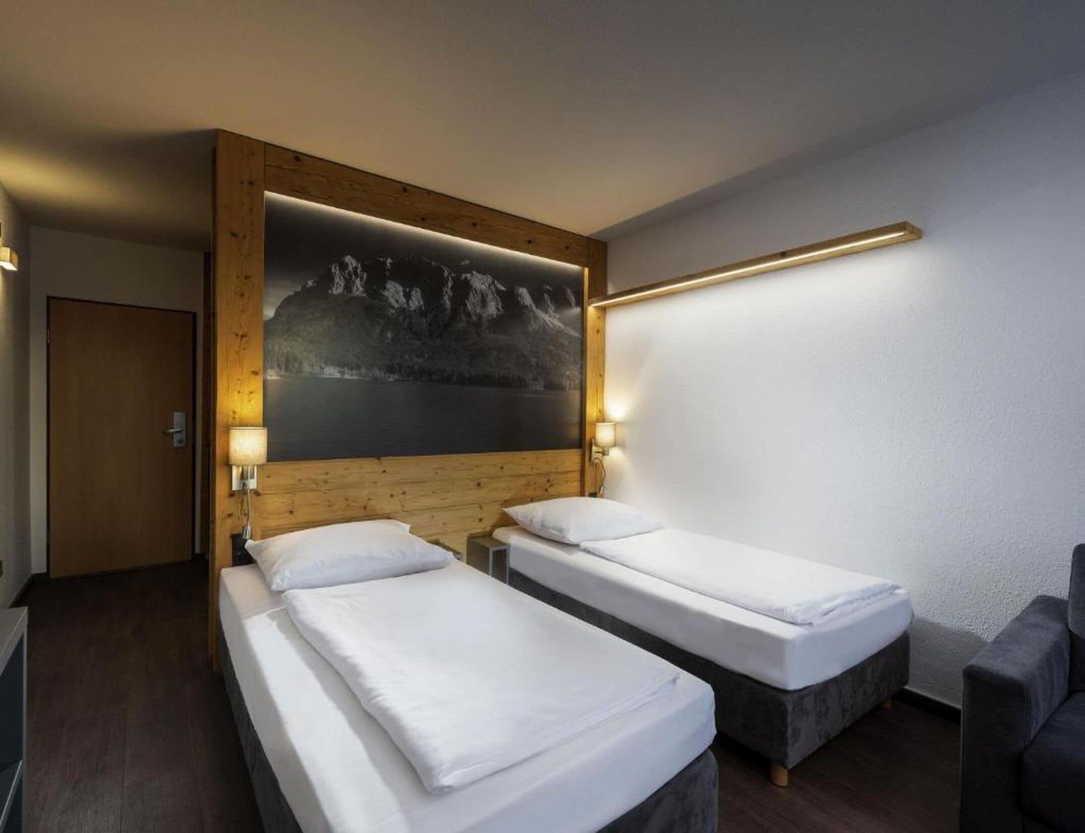 A hotel room with two single beds, a wooden headboard, a large wall-mounted painting of a mountain landscape, and a bedside table with a lamp.