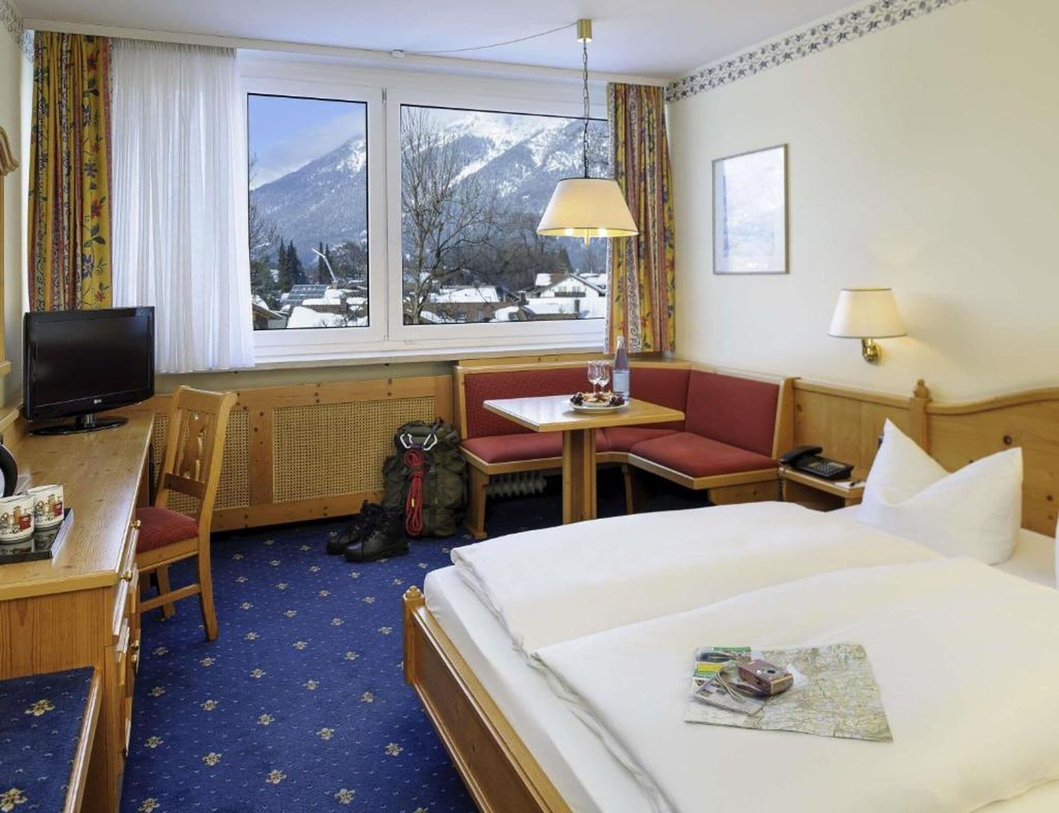 A hotel room with a bed, a desk with a television, a chair, a table with a lamp, and a view of snow-covered mountains through the window.