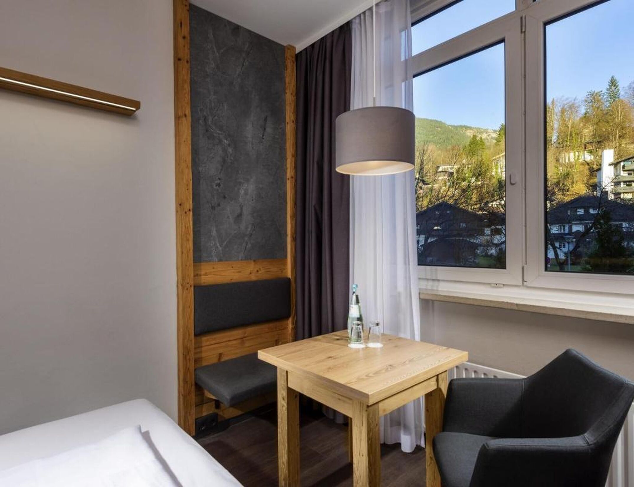Ein Hotelzimmer mit einem Bett, einem Holztisch, einem Stuhl, einer Lampe und einem Fenster mit Blick auf Gebäude und Bäume.