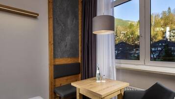 Ein Hotelzimmer mit einem Bett, einem Holztisch, einem Stuhl, einer Lampe und einem Fenster mit Blick auf Gebäude und Bäume.