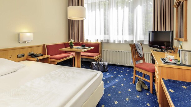 A hotel room with a bed, a desk with a computer, a chair, a sofa, a table with a lamp, a TV, and a window with curtains. - Sunday Hotel Garmisch Partenkirchen (Garmisch-Partenkirchen)