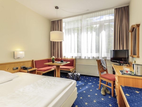 A hotel room with a bed, a desk with a computer, a chair, a sofa, a table with a lamp, a TV, and a window with curtains. - Sunday Hotel Garmisch Partenkirchen (Garmisch-Partenkirchen)