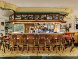 A bar area with wooden bar stools, a counter with various bottles, and framed pictures on the wall. - Sunday Hotel Garmisch Partenkirchen (Garmisch-Partenkirchen)