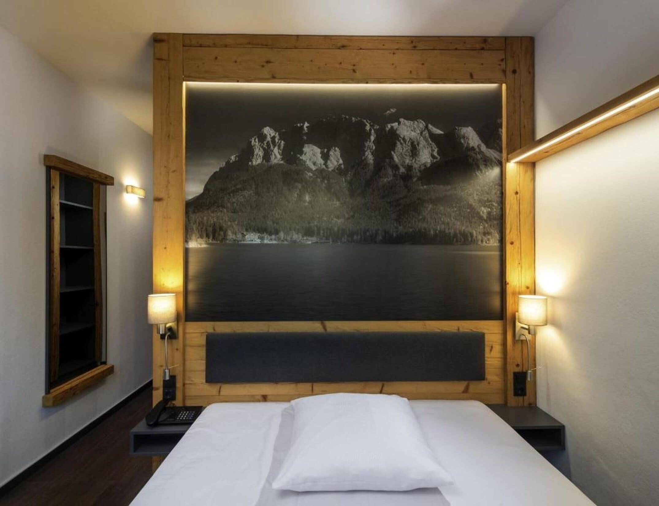 A hotel room with a bed, a nightstand, a telephone, and a wall-mounted painting of a mountain landscape.