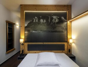 A hotel room with a bed, a nightstand, a telephone, and a wall-mounted painting of a mountain landscape. - Sunday Hotel Garmisch Partenkirchen (Garmisch-Partenkirchen)