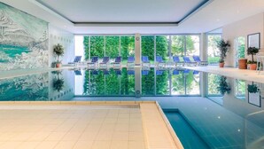 An indoor swimming pool with clear glass walls, blue lounge chairs, and a view of greenery outside. - Sunday Hotel Garmisch Partenkirchen (Garmisch-Partenkirchen)