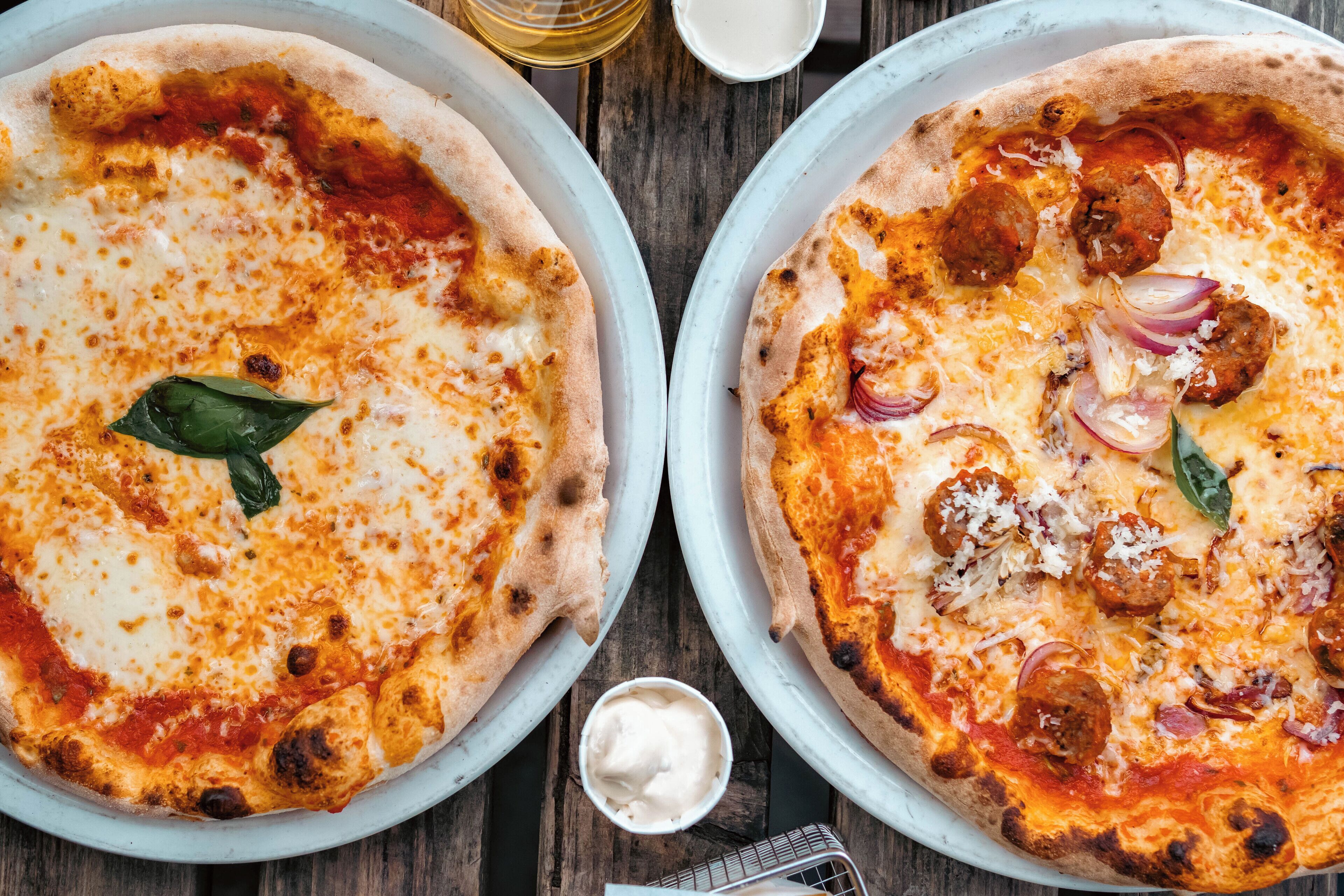 Two pizzas on white pans on a wooden table.