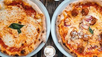 Two pizzas on white pans on a wooden table.
