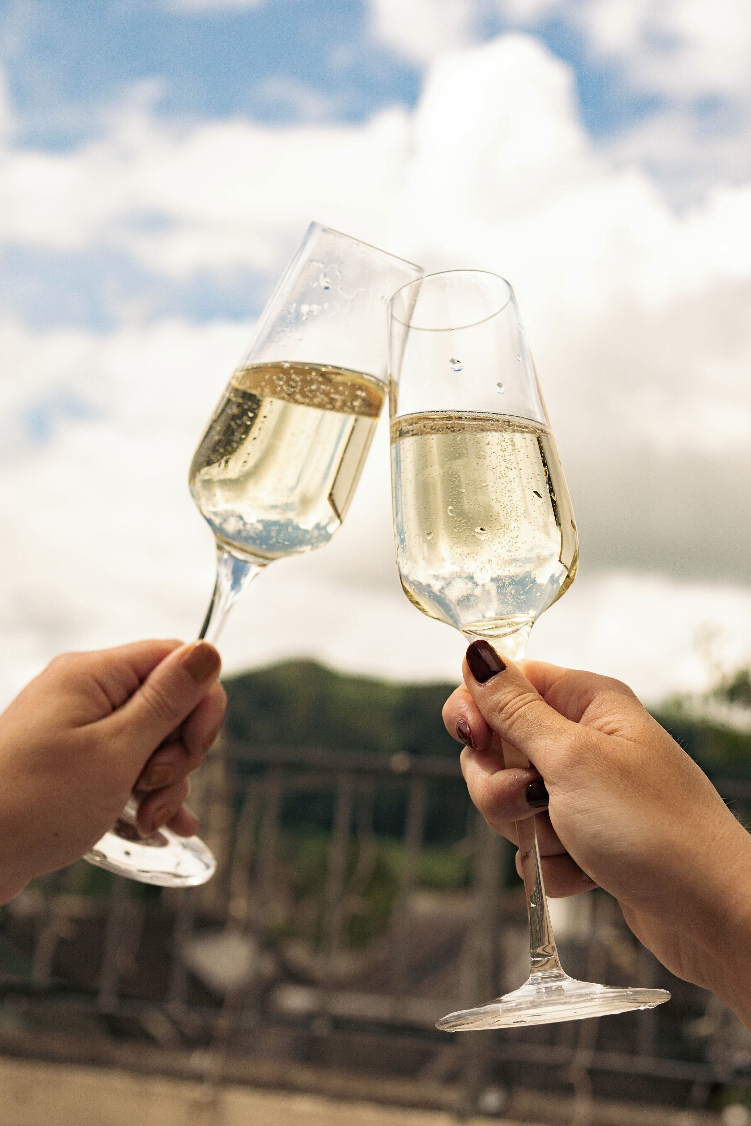 Dos manos sostienen copas de vino blanco sobre un fondo exterior desenfocado.