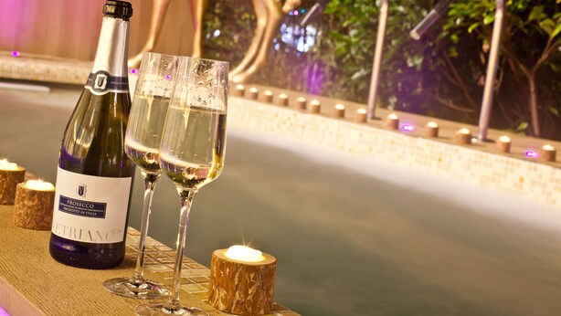 Een flesje Prosecco en twee wijnglazen op een houten tafel, nabij een fontein.