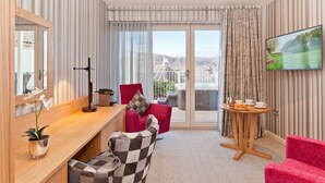 Een moderne hotelkamer met een houten bureau, een rode fauteuil, een geruit bankje, een klein tafeltje met kop en schotel, en een aan de muur bevestigde televisie.