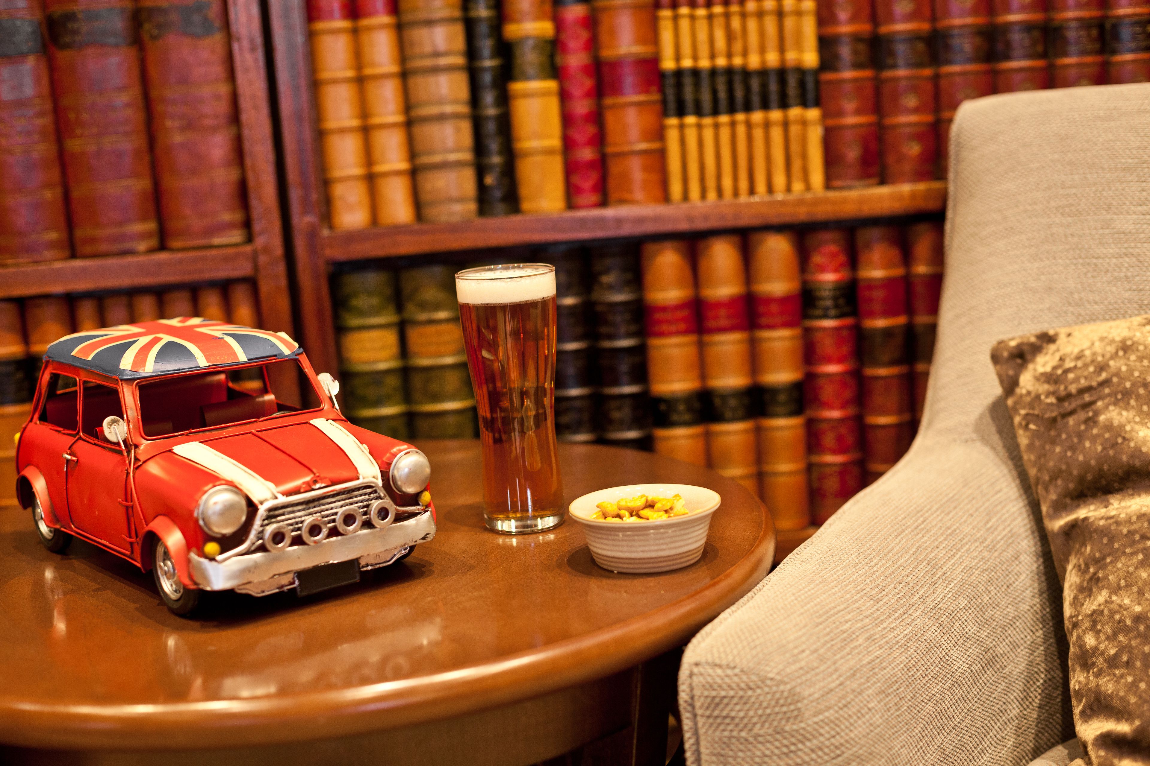 A vintage toy car, a tall glass of beer, and a bowl of corn on the cob on a round wooden table, with a bookshelf in the background.