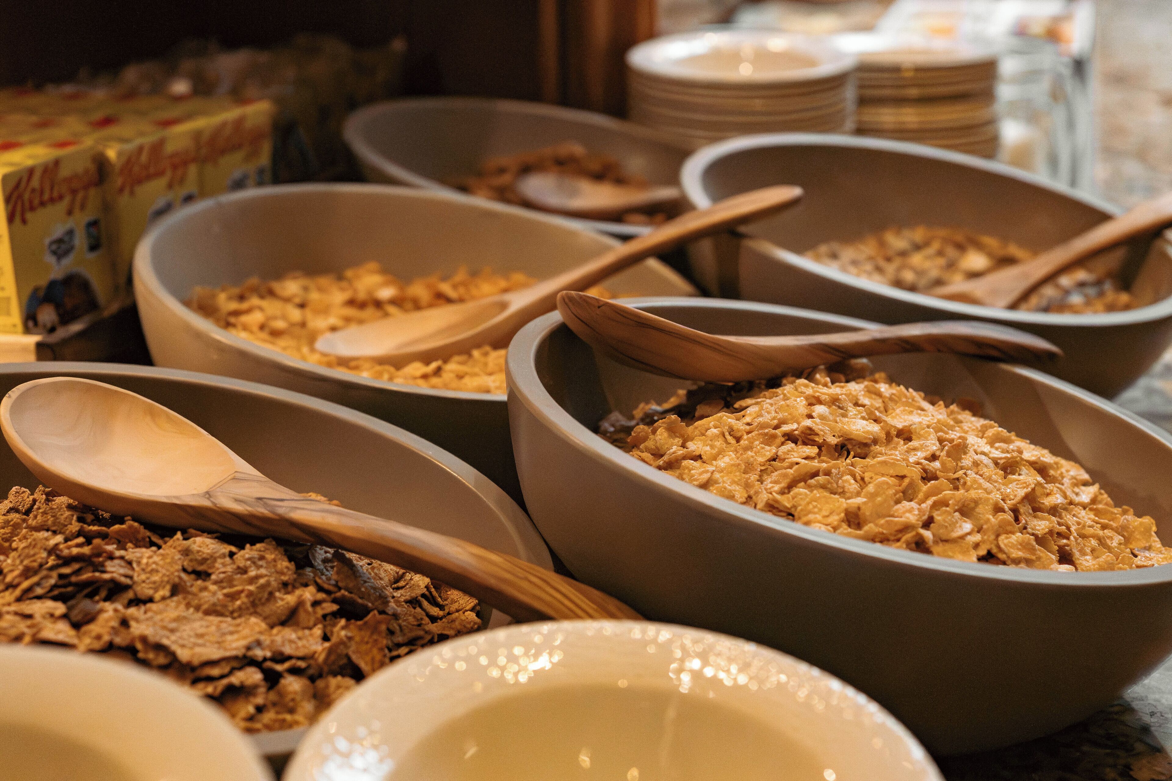 Bowls of cereal with wooden spoons, a box of Kellogg's cereal, and stacked plates in the background.