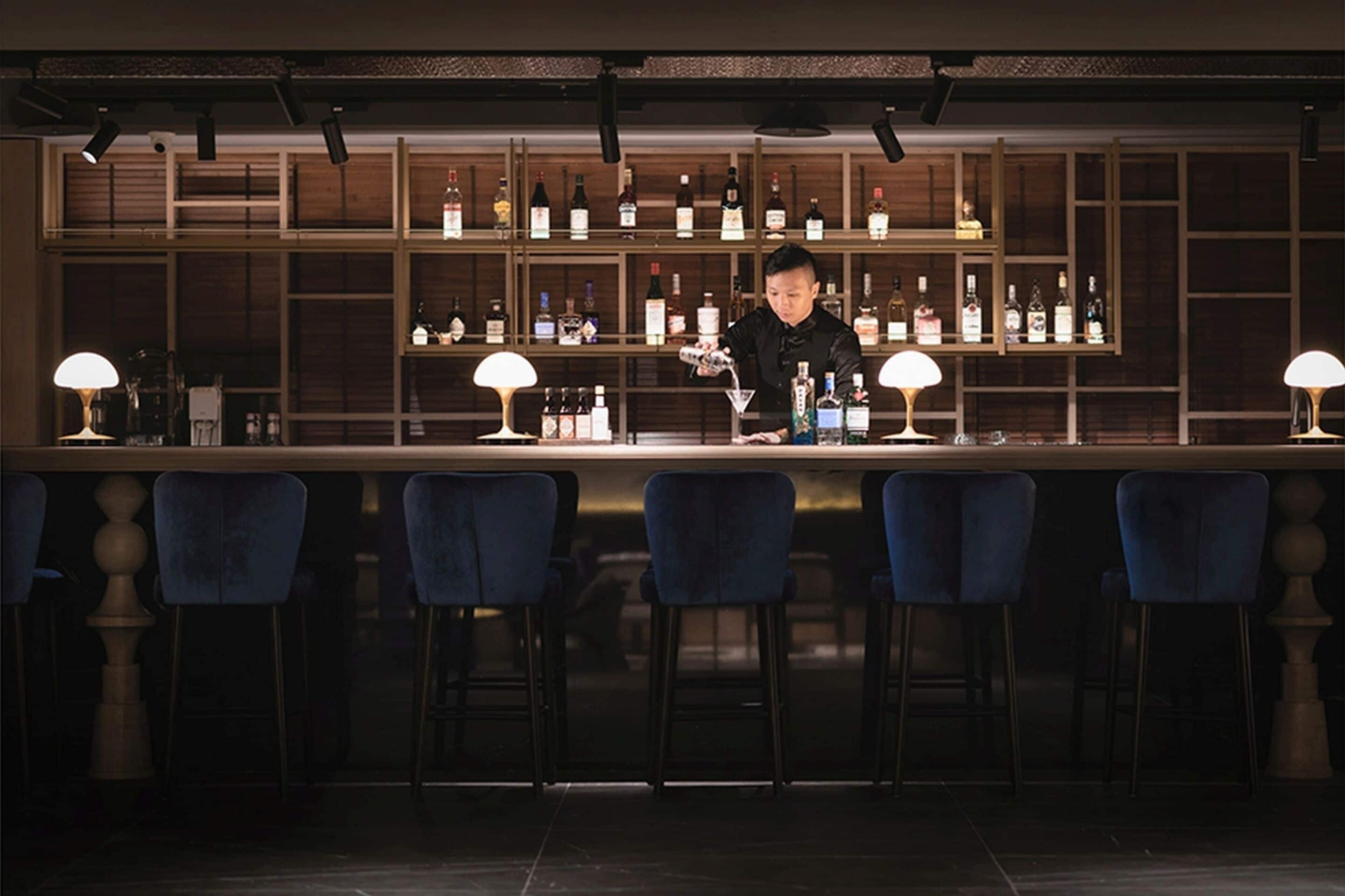 Zona de bar con un cantinero preparando bebidas, taburetes y estanterías llenas de botellas.