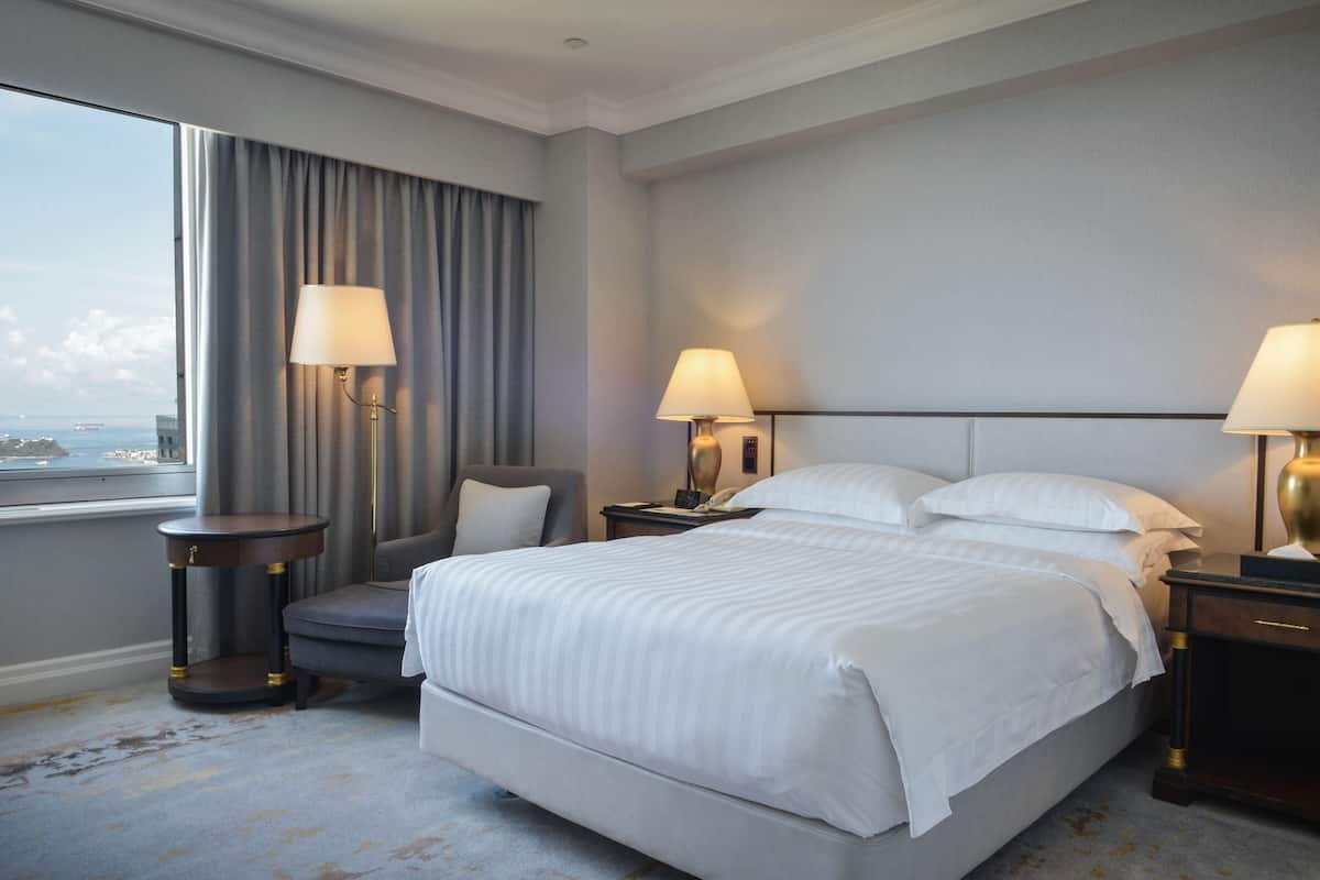 Ein Hotelzimmer mit einem großen Bett, zwei Nachttischen mit Lampen, einem Sessel und Blick auf die Stadt durch ein Fenster.