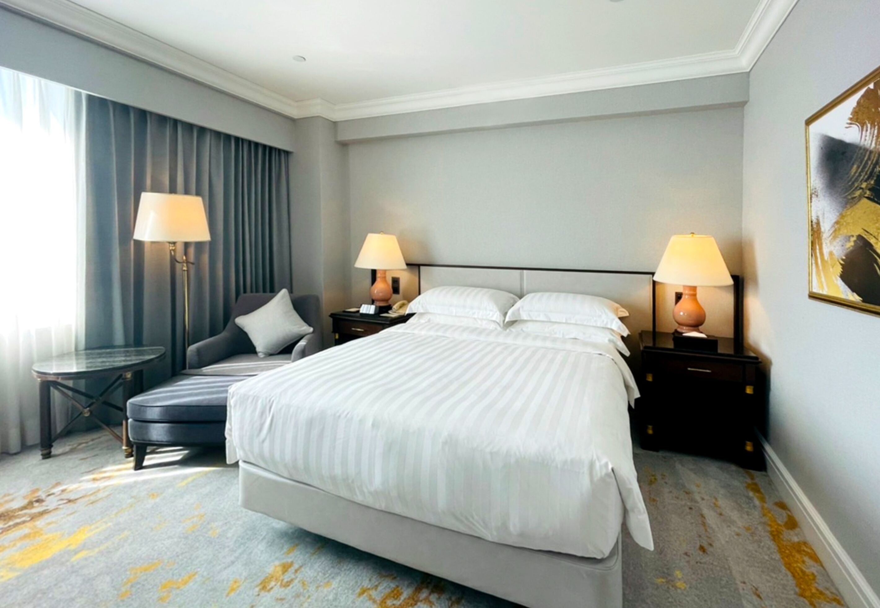 Una habitación de hotel con una cama grande, dos mesitas de noche con lámparas, un sofá pequeño y un ventanal con cortinas.