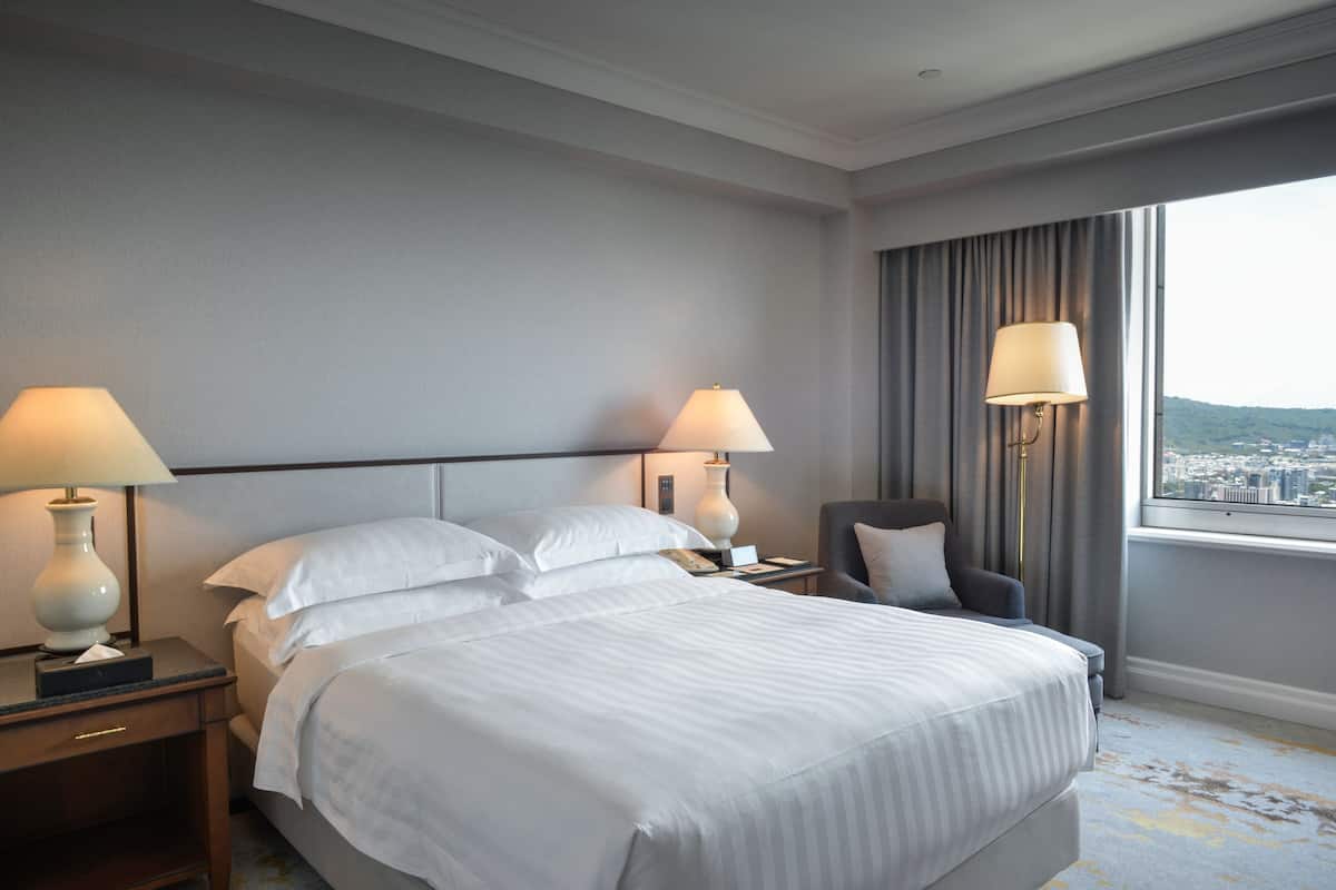 Ein Hotelzimmer mit einem großen Bett, zwei Nachttischen mit Lampen, einem Sessel und Blick auf eine Stadtkulisse durch ein großes Fenster.