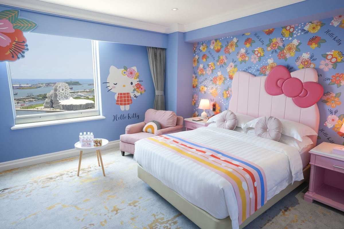 Ein Schlafzimmer mit einem großen Bett, einem Fenster mit Blick auf eine Stadtansicht, einer Hello Kitty Wandsticker und einem floralen Tapetenmuster.