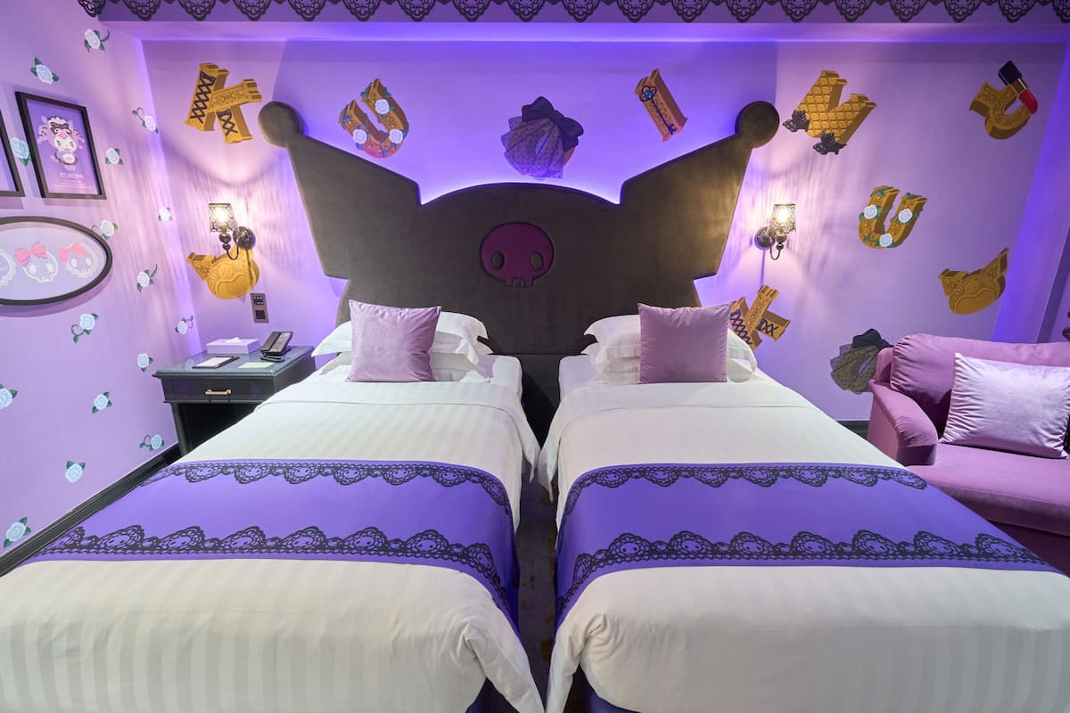 Ein Zimmer mit zwei Betten, violetter Bettwäsche, einem Totopfkopf als Kopfteil und Wanddekorationen, darunter Buchstaben sowie ein Hello Kitty Schild.