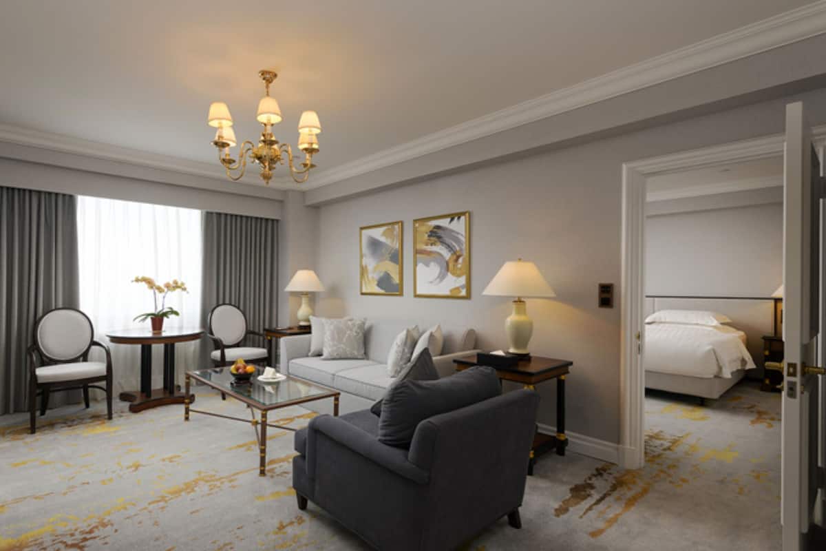 Ein Hotelzimmer mit grauem Sofa, rundem Couchtisch, Fernseher, Bett und einer Chandelier.