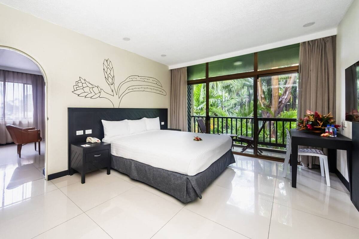 A hotel room with a bed, a desk, a chair, a nightstand, and a view of greenery through large windows.