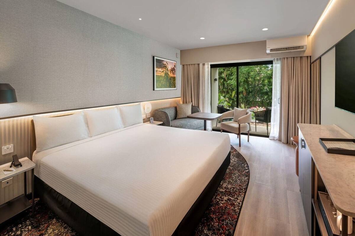 A hotel room with a large bed, a desk, a chair, a sofa, and a view of greenery outside.