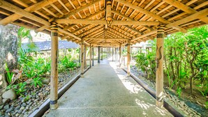 A covered walkway with wooden beams and columns, surrounded by greenery and a stone pathway. - Tanoa International Hotel (Nadi)
