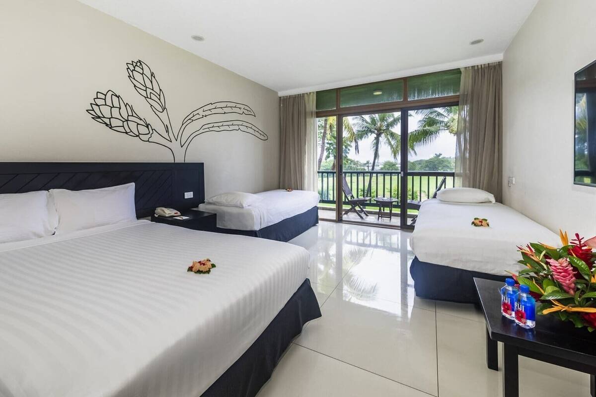 A hotel room with two beds, a balcony with a view of palm trees, and a table with a floral arrangement and water bottles.